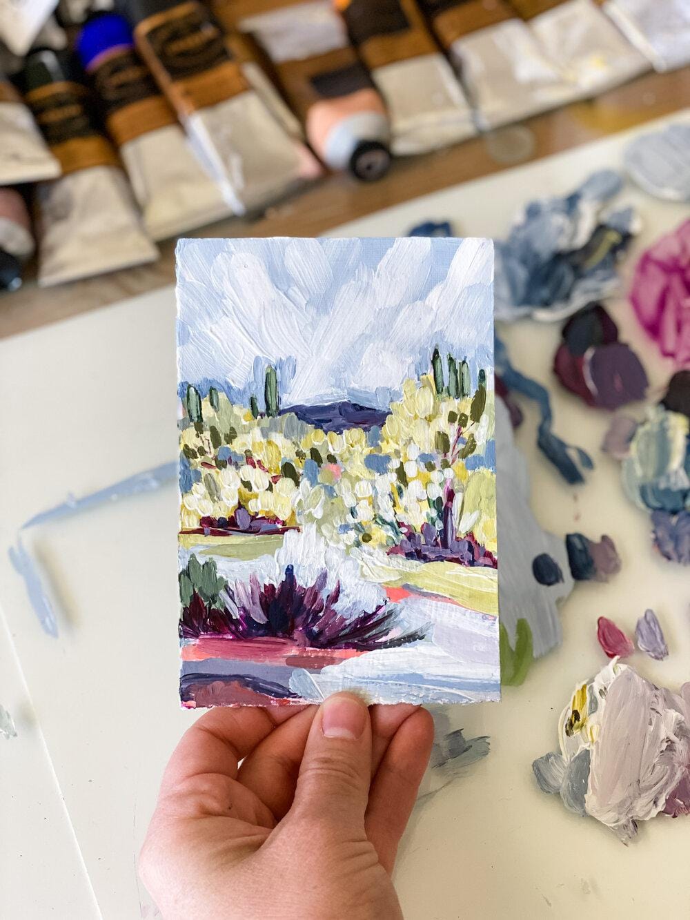 Hand holding a small landscape painting with paint tubes in background.