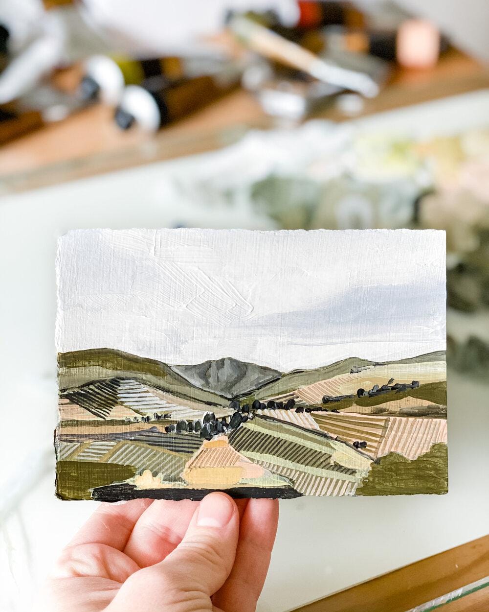 Hand holding a small landscape painting of rolling hills.