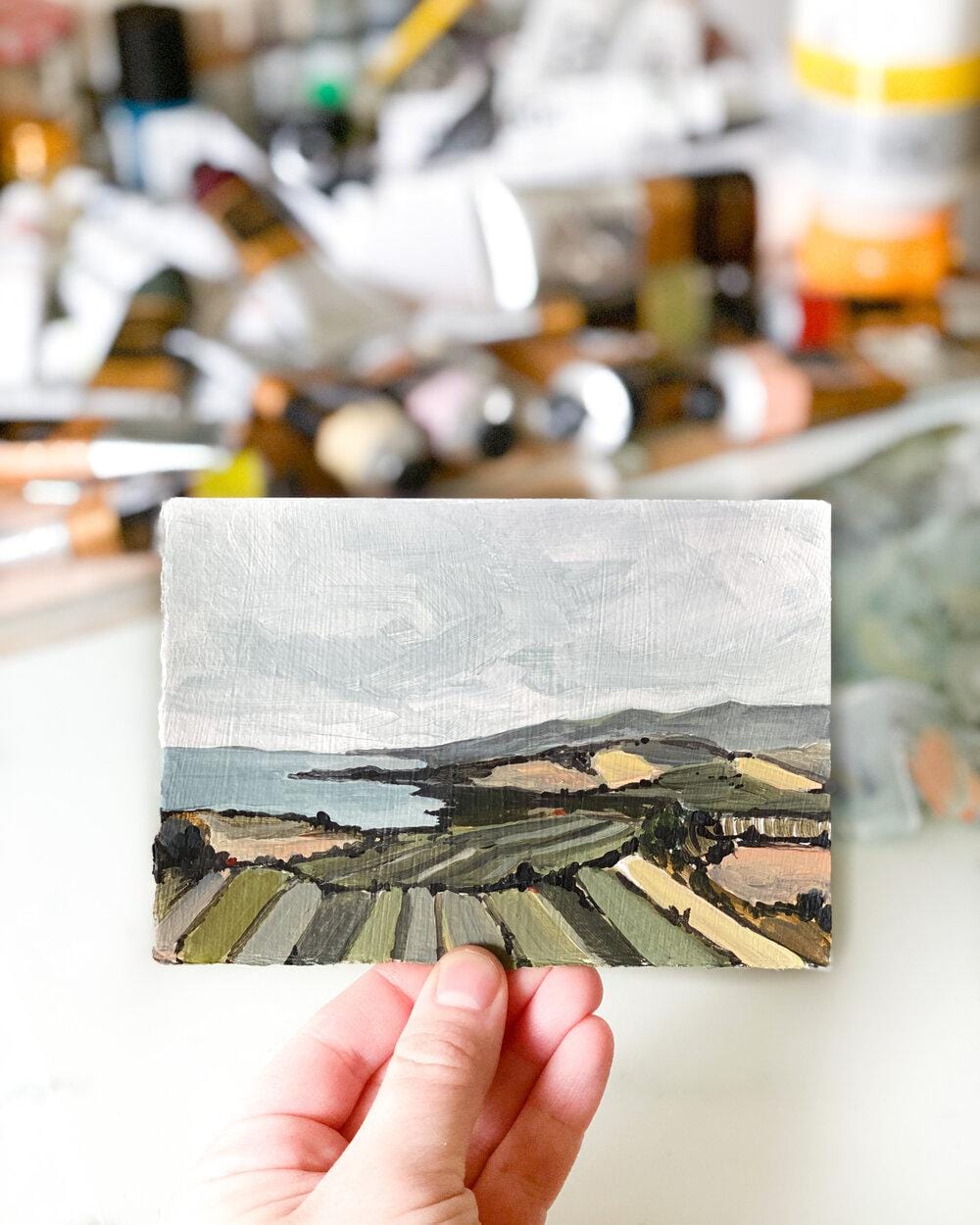 Hand holding a landscape painting of fields and ocean.