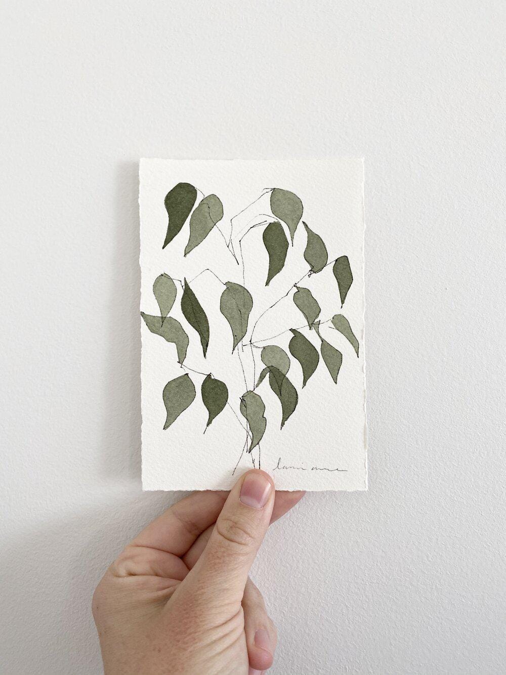 Hand holding a watercolor painting of green leaves on a white background.