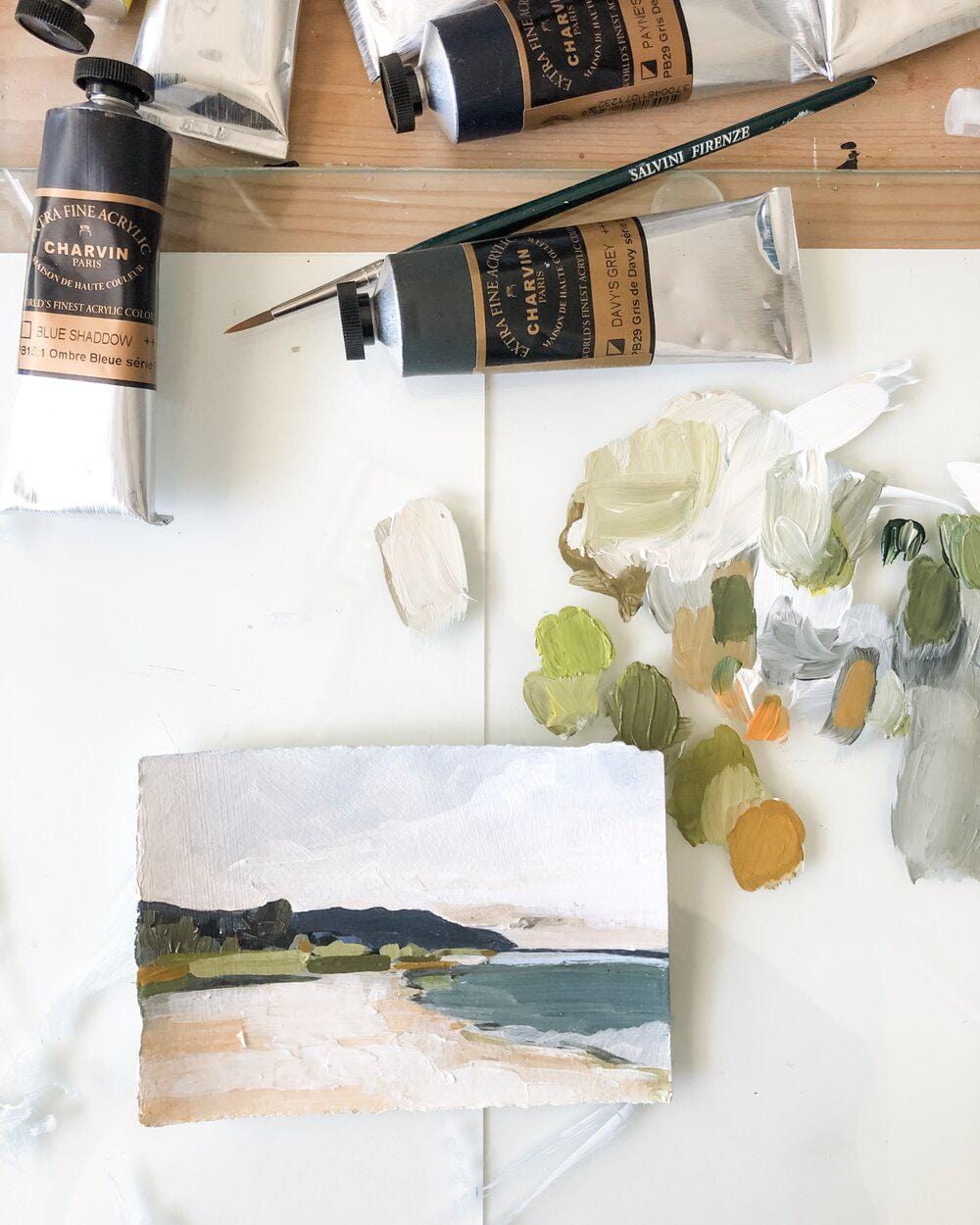 Paint tubes, brush, and landscape painting on a white surface.