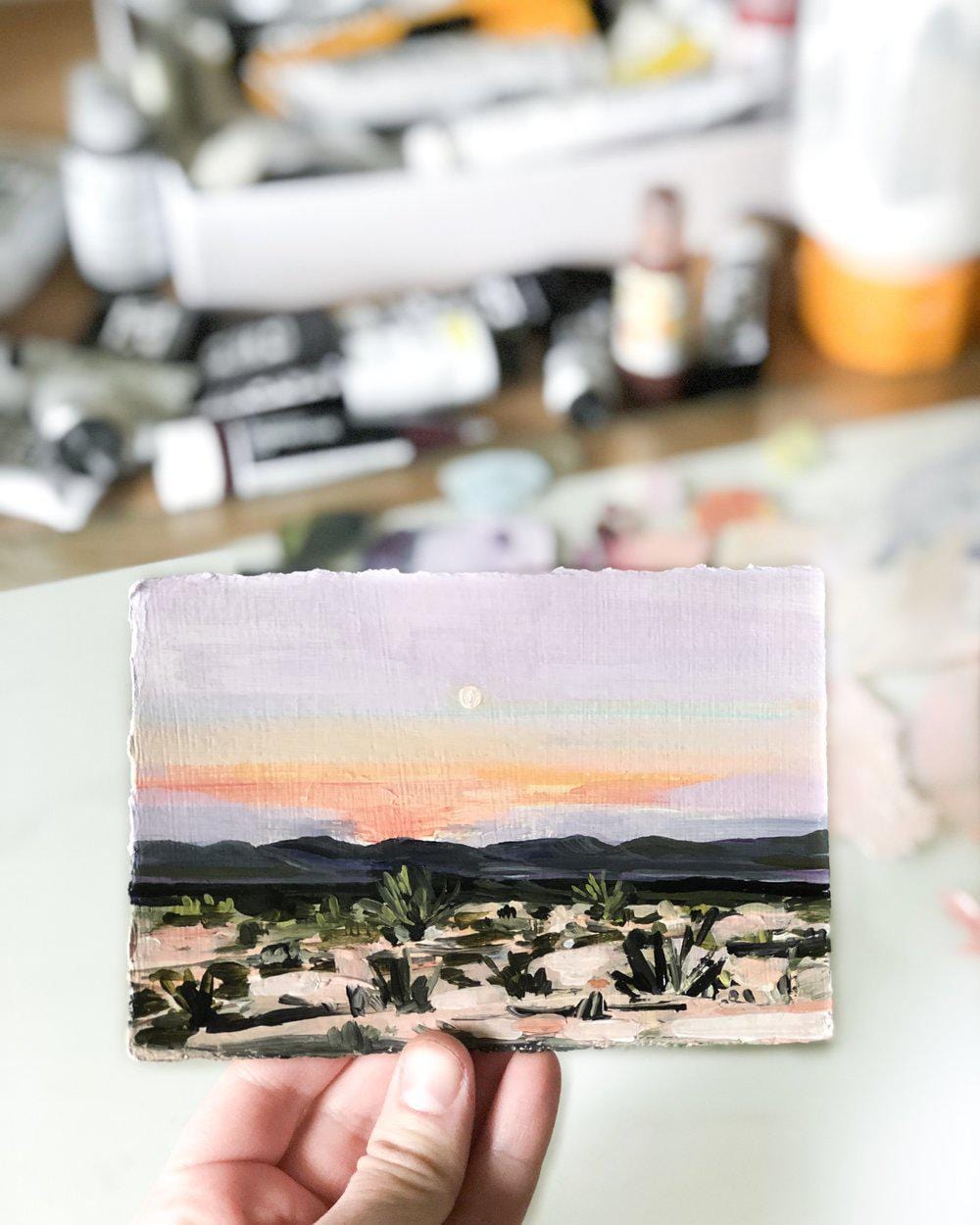 Hand holding small landscape painting of sunset.