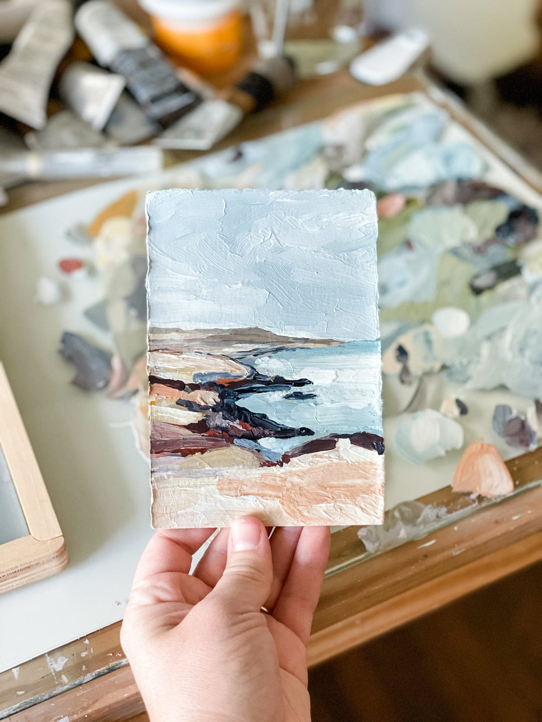 Hand holding a small landscape painting of a beach.