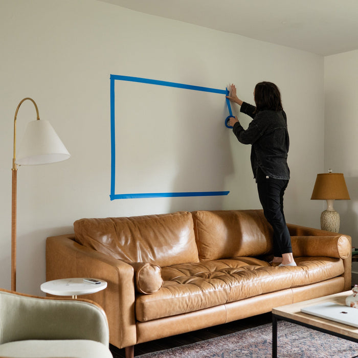 a woman using painters tape to outline the size of art over a sofa