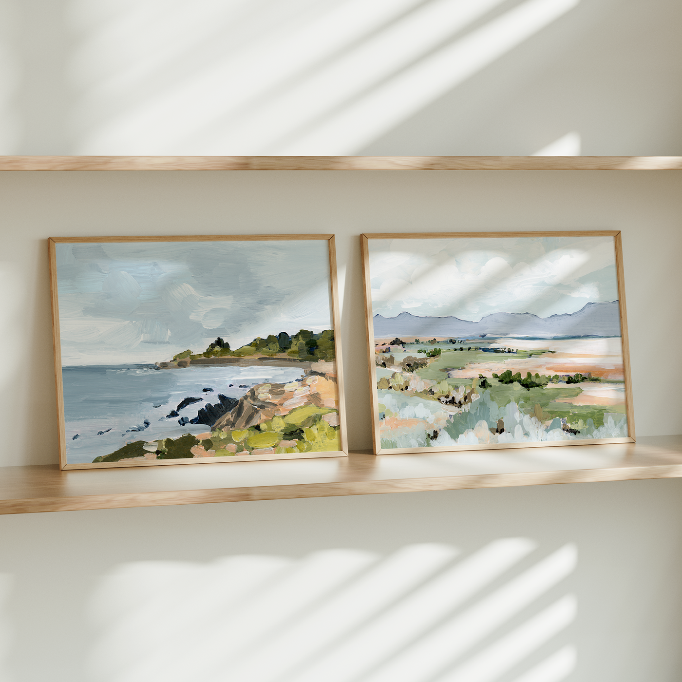 Two framed landscape paintings on a wooden shelf with sunlight casting shadows.