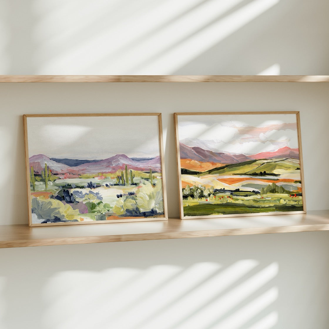 Two framed landscape paintings on a wooden shelf with sunlight casting shadows.