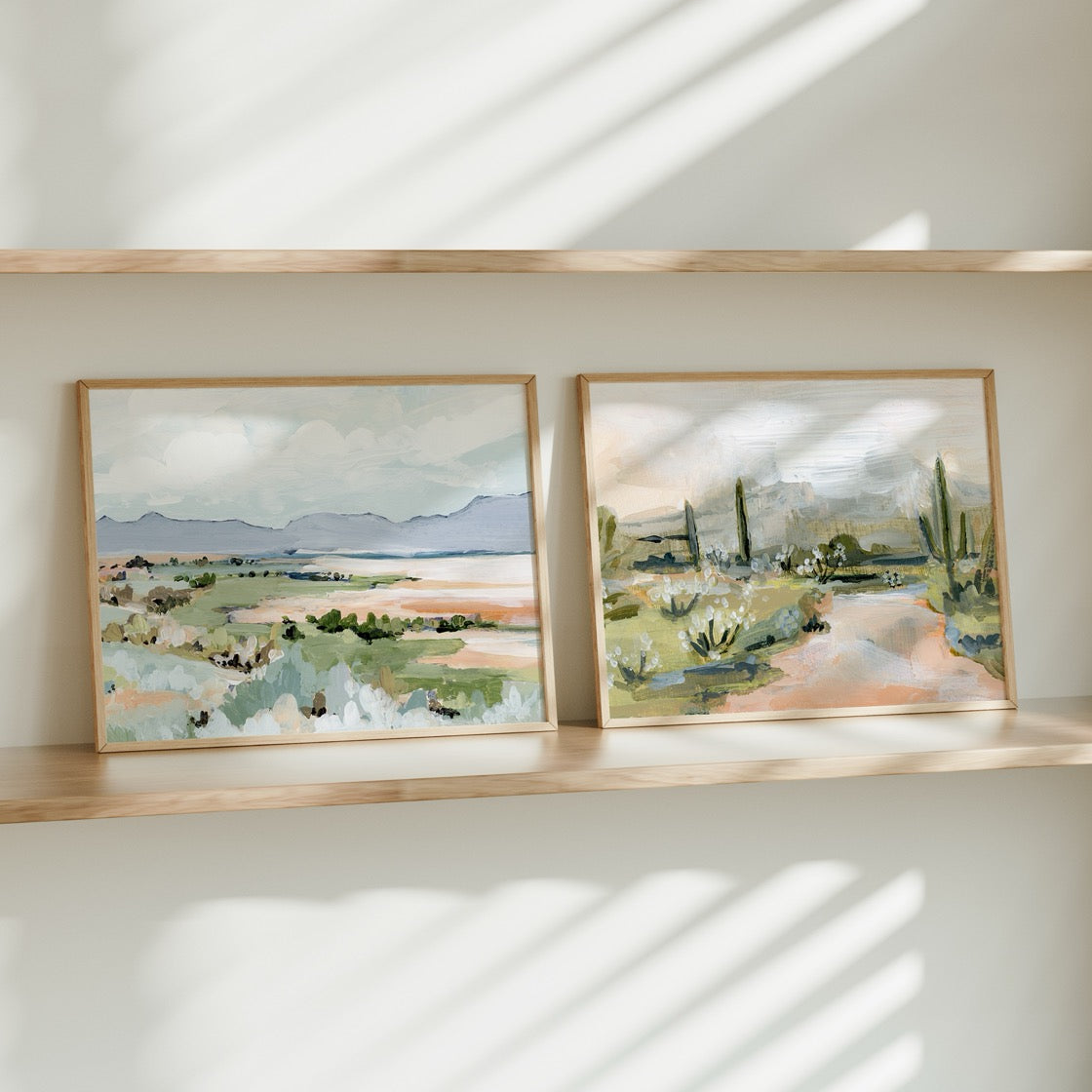 Two framed landscape paintings on a wooden shelf with sunlight casting shadows.