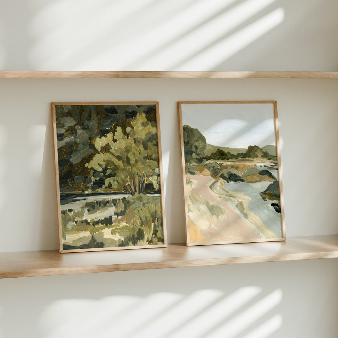 Two framed landscape paintings on a wooden shelf with sunlight casting shadows.